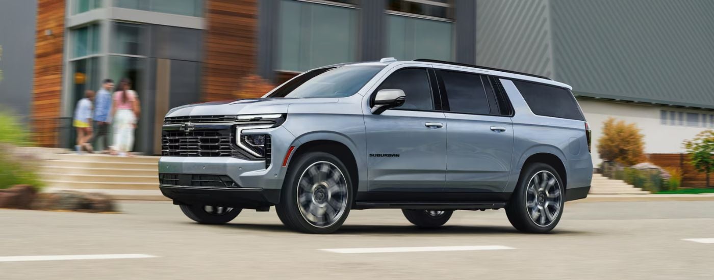 Silver 2026 Chevy Suburban driving by a building.